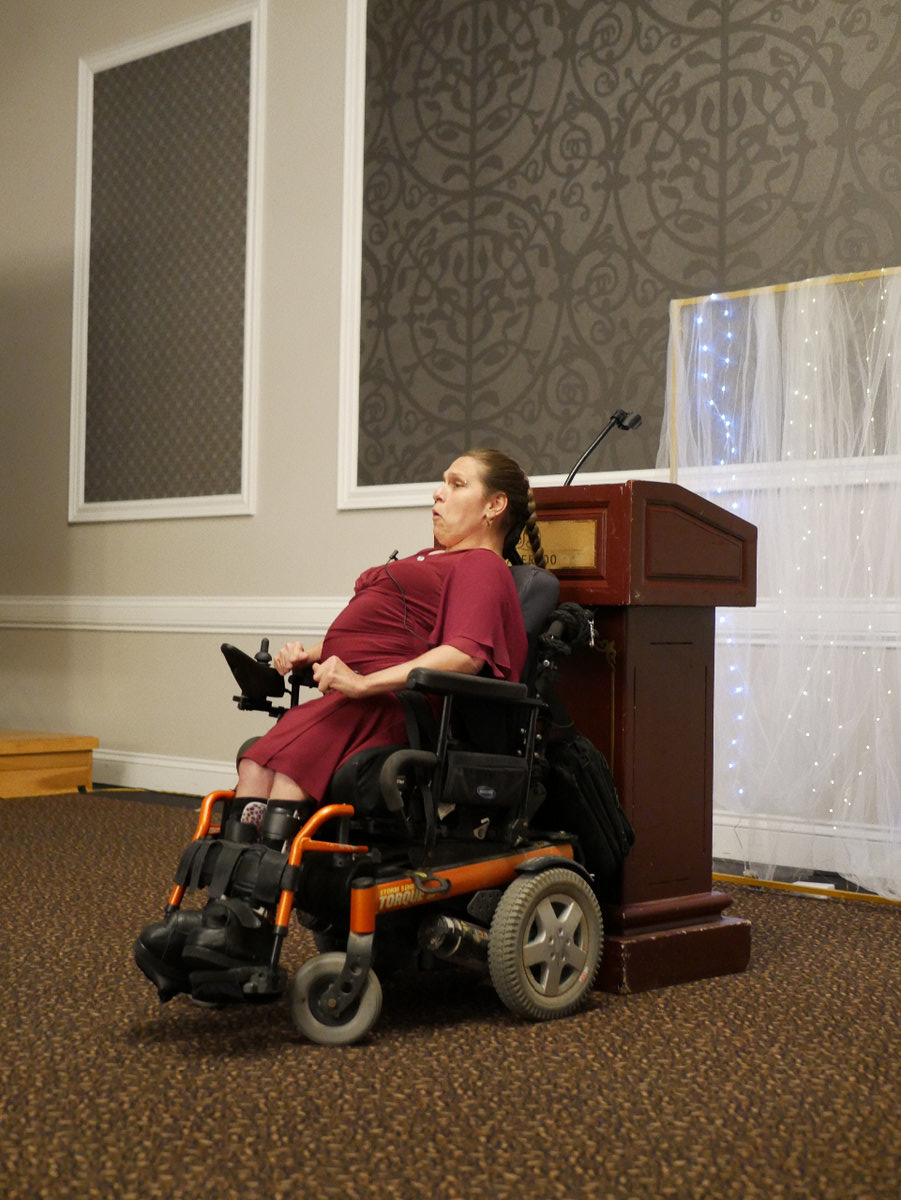 A woman in a purple dress uses an electric wheelchair with orange-coloured metal parts. Her wheelchair is positioned in front of a podium. She is mid-sentence in her speech.